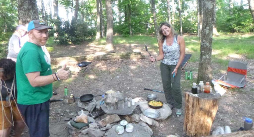 Two people prepare food in a wooded area. 